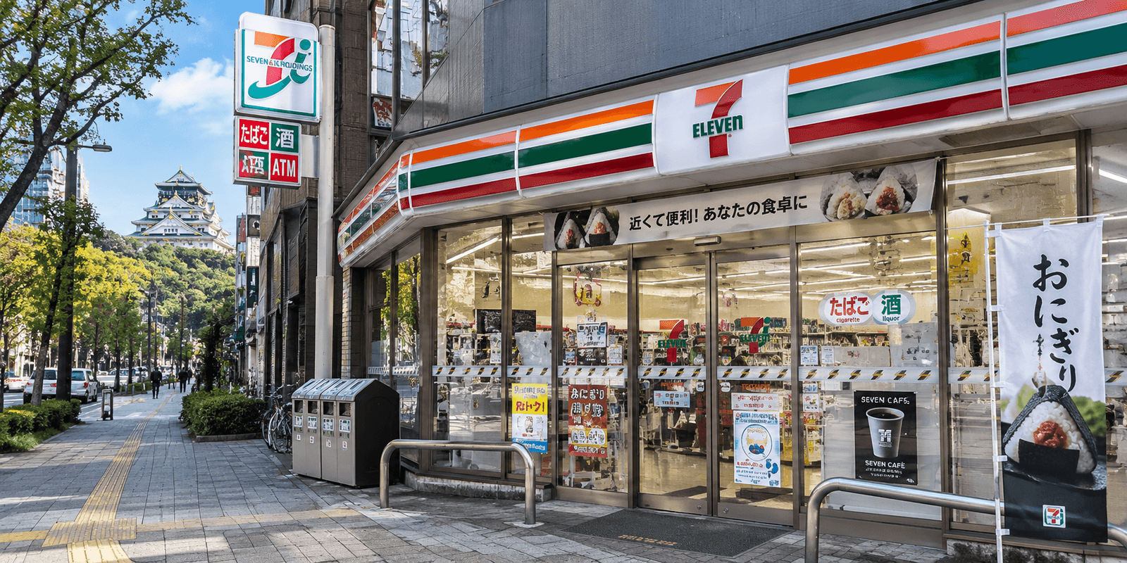 A 7-Eleven convenience store storefront on a tree-lined street in Osaka, Japan, with Osaka Castle visible in the distance