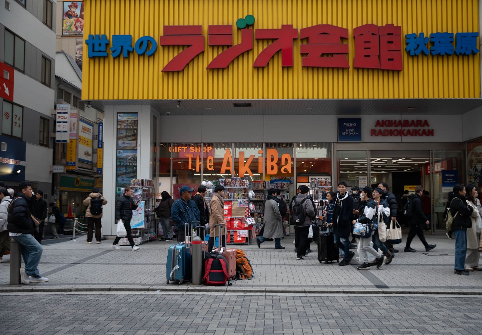 Travelers' suitcases and backpacks gathered on the pavement outside Akihabara Radio Kaikan in Tokyo while shoppers walk past