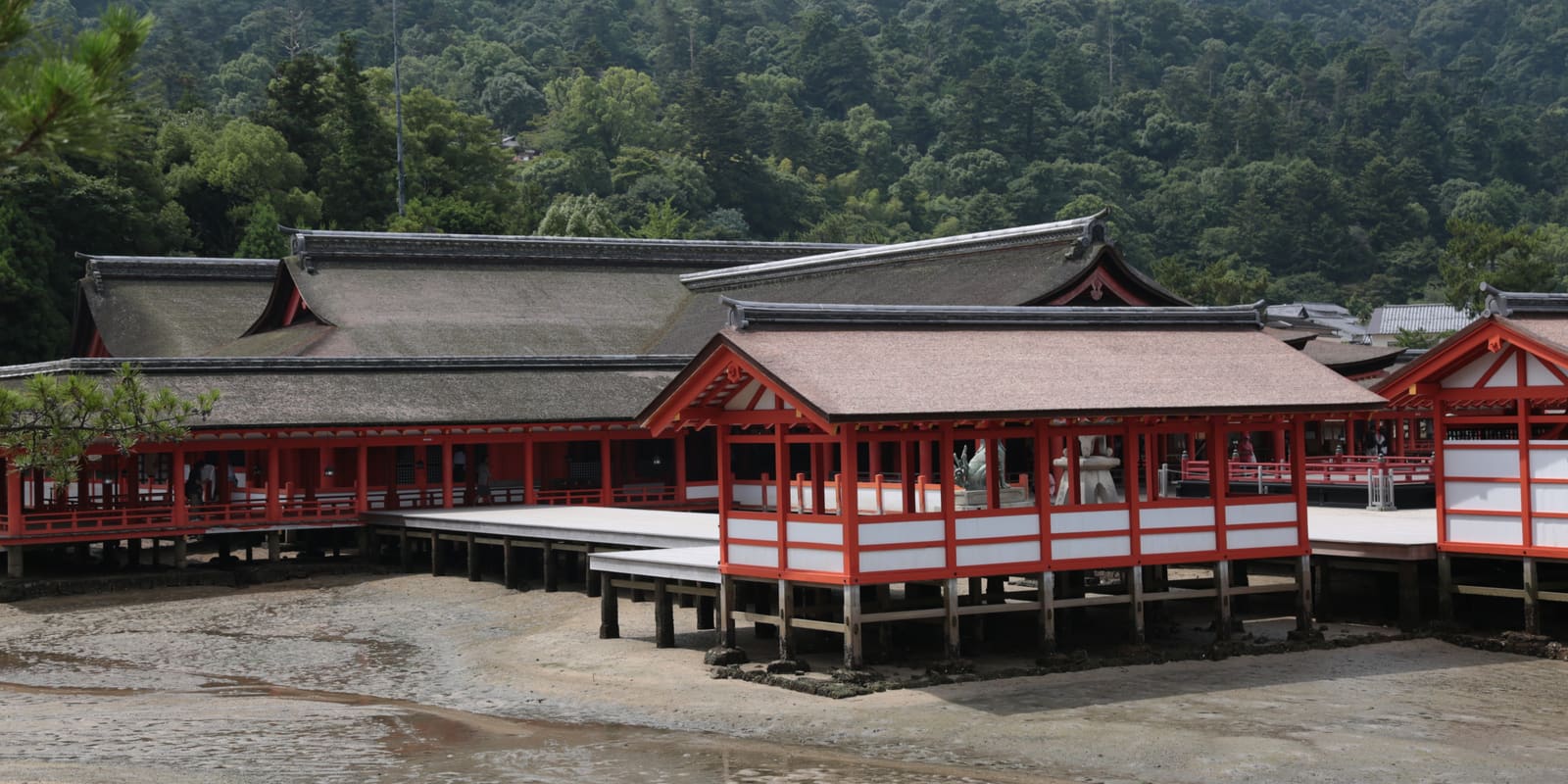 Vermilion halls and raised corridors of Itsukushima Shrine at low tide on Miyajima, with forested slopes behind