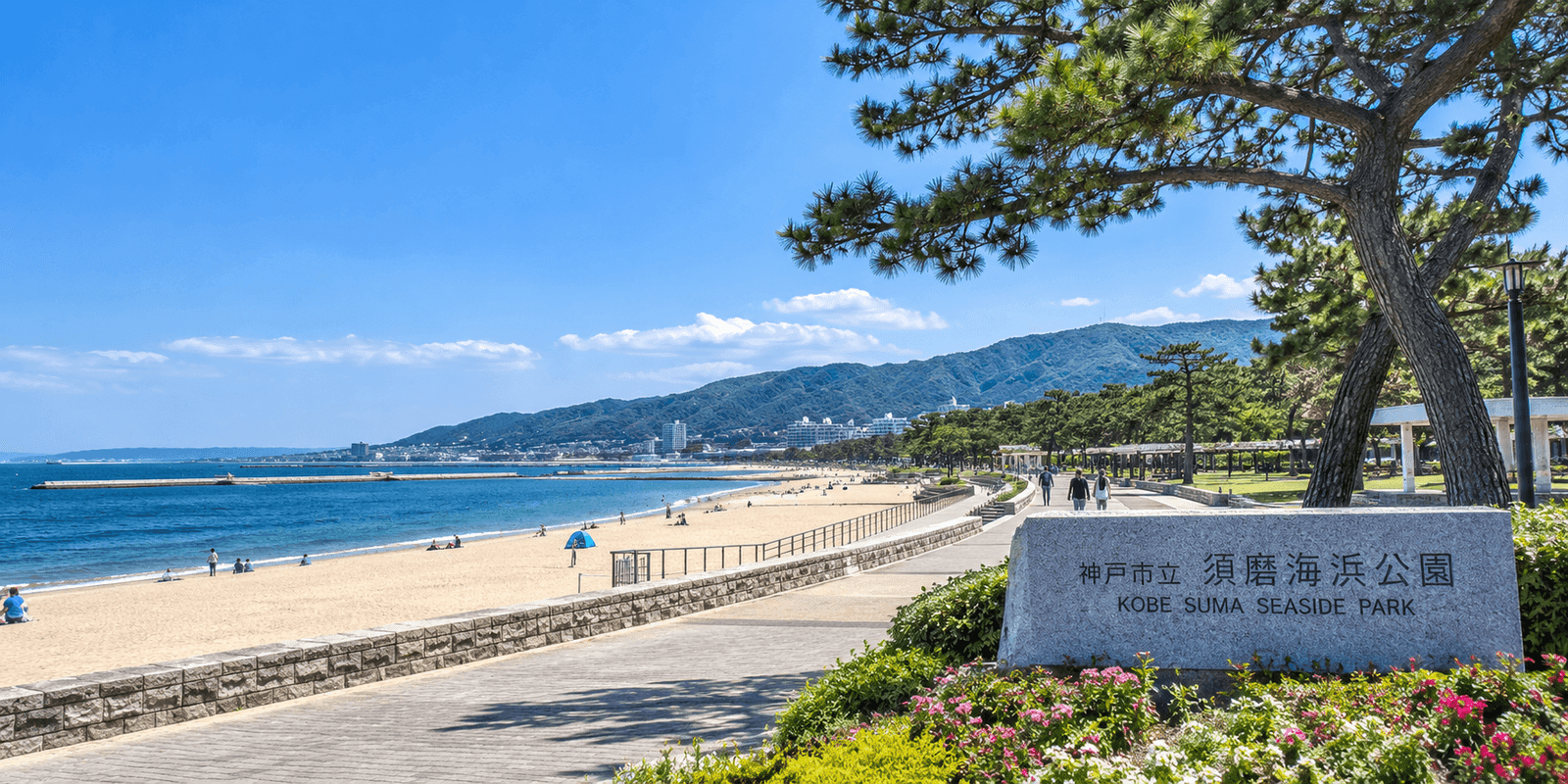 Sandy beach and waterfront promenade at Kobe Suma Seaside Park with the park's stone entrance marker in the foreground
