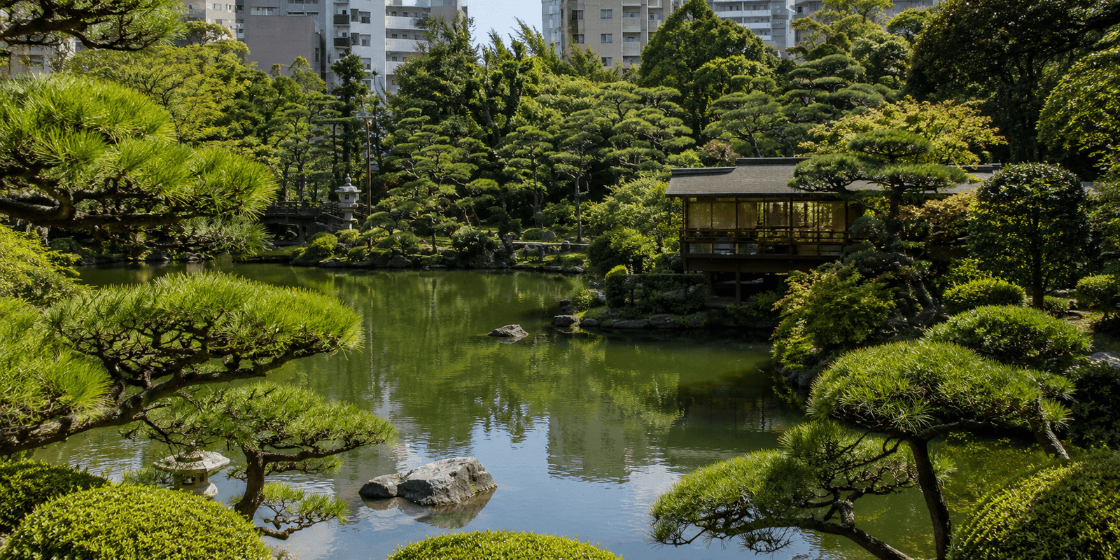 Pond, pine trees, and a lakeside teahouse-style building at Sorakuen Garden with Kobe high-rises visible behind