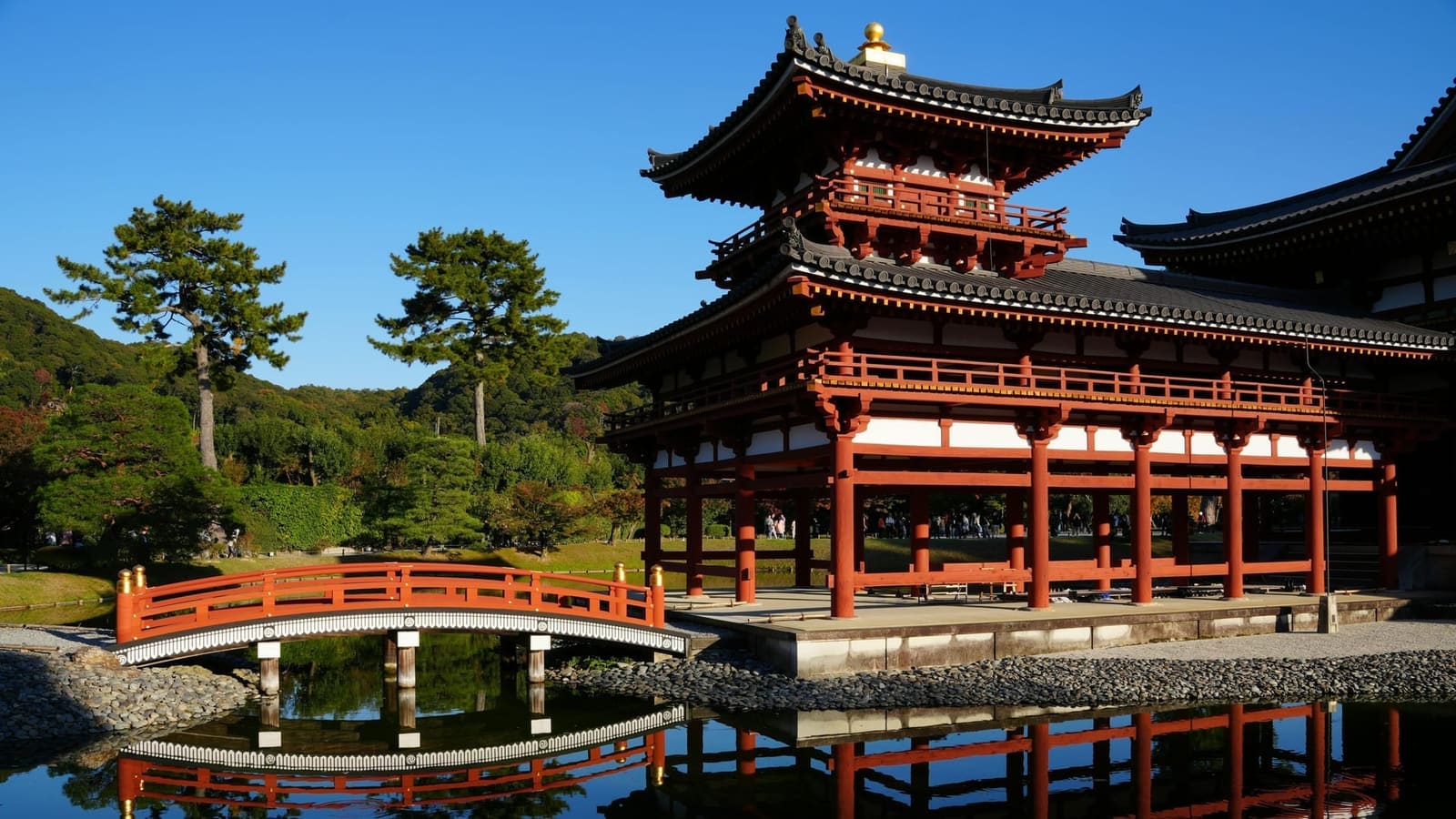 A corner tower of Byodo-in's Phoenix Hall and a vermilion bridge reflected in the pond under a clear autumn sky