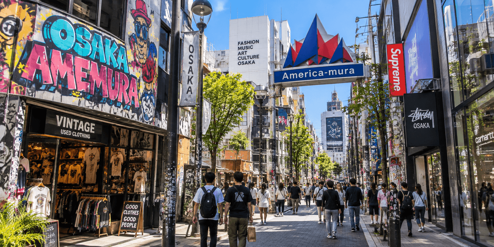 Amerikamura street in Osaka with the blue America-mura gateway sign, a graffiti mural reading Osaka Amemura above a vintage clothing shop, and pedestrians walking under street trees, Japan
