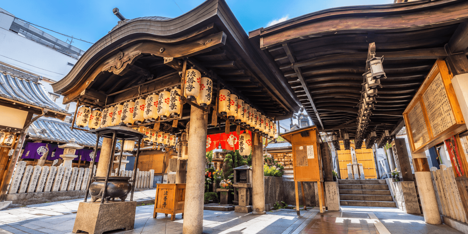 Hozenji Temple's wooden canopy hung with paper lanterns marked with the temple's name in Japanese, above an incense burner, notice boards, and stone steps in the precinct, Namba, Osaka