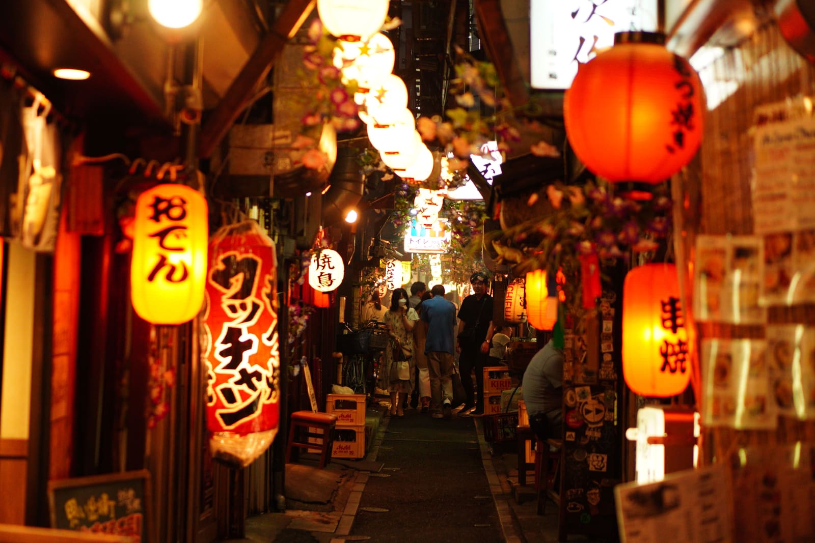 Narrow alley at night lined with small eateries, glowing paper lanterns for yakitori, oden, and skewers, stacked beer crates, and diners walking toward the far end