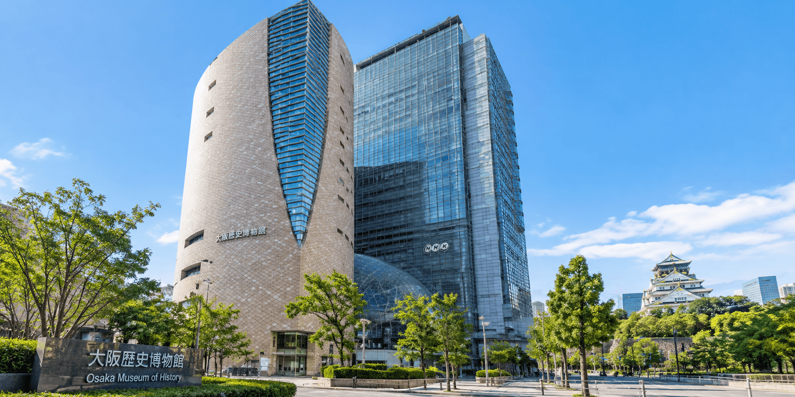 Osaka Museum of History's curved modern exterior with its own Japanese and English signage, adjoining the NHK Osaka tower, with Osaka Castle visible in the background, Osaka Japan
