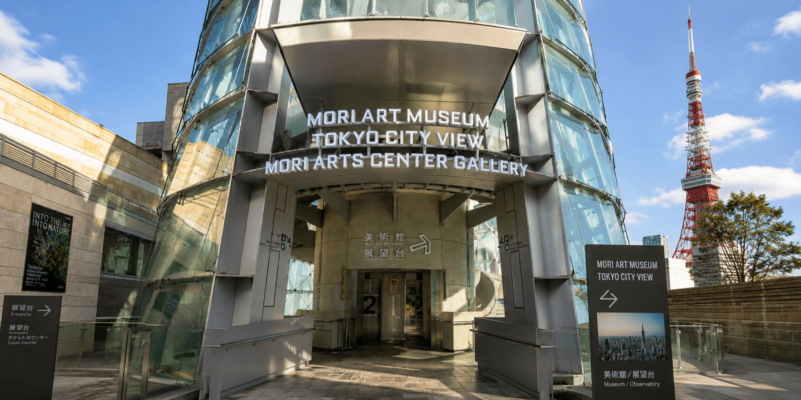 Mori Art Museum entrance signage atop Mori Tower, with Tokyo City View and Mori Arts Center Gallery signage alongside it and Tokyo Tower visible through the glass