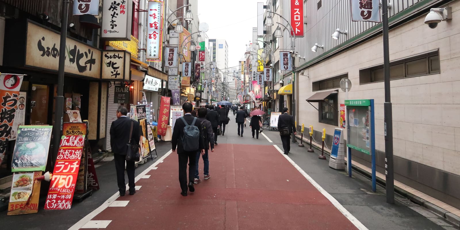 A restaurant-lined side street in Ikebukuro with ramen shop signs and office workers walking